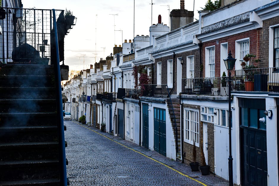 A row of terraced brick houses with white-painted facades, small front gardens, and black metal railings along the pavement in West Hampstead, captured during early evening. The front doors are predominantly painted in dark shades, and some houses have potted plants or small decorative items on the balconies or window sills. The narrow cobbled street is lined with a yellow line along the curb, indicating parking restrictions. Warm light from the setting sun briefly illuminates parts of the street, with shadows cast on the buildings. On the left side, a dark external staircase with metal railings ascends to an upper level, possibly leading to a basement or side entrance, and is partially visible in the foreground. The scene reflects a typical residential area suitable for home relocation or furniture transport services, like those provided by Man and Van West Hampstead, with the street prepared for moving logistics, such as loading or unloading furniture during a house removal.
