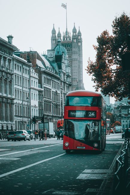 A red double-decker bus designated with route number 24 is parked on a busy city street in West Hampstead, London, with historic buildings lining the road and the iconic Tower of London visible in the background. The scene includes several pedestrians walking on the sidewalk, some waiting at a bus stop, while cars are driving past. On the right side of the street, a man and a woman are seen carrying cardboard boxes and plastic-wrapped furniture, likely during a home relocation or moving process. Nearby, a small trolley is used for transporting packing materials and smaller boxes, with additional packing supplies visible near the curb. This setting illustrates urban logistics related to house removals, with the busy street environment emphasizing the importance of careful planning in furniture transport and packing for local moves. The natural lighting suggests daytime, and the overall scene is calm, reflecting a typical move happening within the city street environment managed by Man and Van West Hampstead.