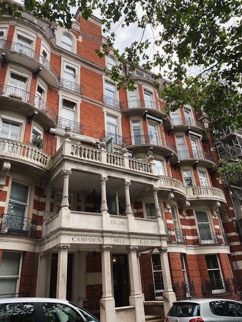 A multi-storey red brick building with white decorative stone details and ornate balconies, located on Camden Hill in West Hampstead. The building’s entrance features classical columns supporting a balcony, with the address sign '13-36 Camden Hill Court' above the doorway. Tree branches with green leaves partially obscure the upper floors and cast shade on the facade. Parked cars are visible on the street in front of the building, and the overall scene captures the exterior view of a residential property suitable for house removals or furniture transport as part of a home relocation process, relevant to Camden Council moving permit requirements and services offered by Man and Van West Hampstead.
