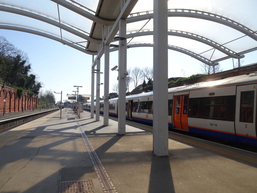 A modern commuter train with a sleek, aerodynamic design is parked at West Hampstead Thameslink station platform. The train's exterior is primarily silver with red and black accents, and its front fea