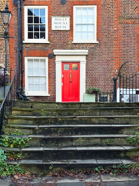 The image shows the front entrance of a traditional brick house with a narrow, steep staircase leading up to a red-painted wooden door. The house features two upper windows with white frames, one on each side of the door, and a sign reading 'Holly Mount House' mounted on the brick wall above the door. The staircase consists of several weathered stone steps with moss and small plants growing in the gaps, flanked by black metal railings on both sides. To the right of the stairs, there is a small green plant in a rectangular planter, and on the left, a black lamp post is attached to the brick wall. The scene captures a typical residential exterior, suggesting a narrow stairway access common in West Hampstead flats, relevant to house removals and furniture transport during home relocation processes facilitated by Man and Van West Hampstead.