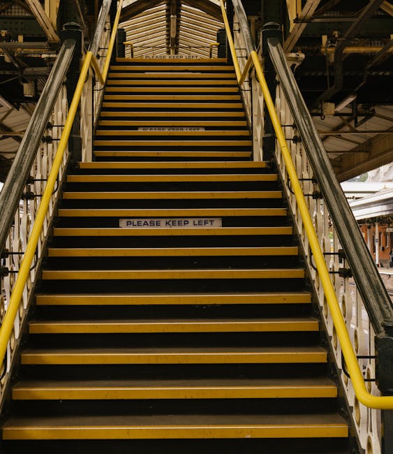 Image of a steep, narrow staircase inside a building used for home relocation and furniture transport, showing black and yellow non-slip treads on each step. The staircase has yellow safety handrails on both sides and is enclosed by metal support beams and a ceiling structure above. Two signs with the text 'PLEASE KEEP LEFT' are visible on the risers. The staircase leads upward toward an illuminated upper level, with some light visible through gaps in the structure. The environment appears to be part of an industrial or multi-storey property, and the clear visual cues support the process of packing, loading, and moving furniture during a house removal service provided by Man and Van West Hampstead.