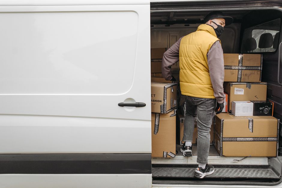 A man wearing a yellow sleeveless jacket, grey trousers, and black and white sneakers is inside the back of a van, which has its sliding door open, revealing a variety of packed cardboard boxes secured with black and white striped tape. The boxes are stacked neatly inside, some containing household items and a small appliance visible on top. The van is parked on pavement outside a property, and the man appears to be in the process of loading or organizing the boxes for a home relocation or furniture transport. The environment is well-lit, indicating daytime, and the focus is on the loading process during a furniture or household move, highlighting packing and moving logistics as managed by Man and Van West Hampstead.