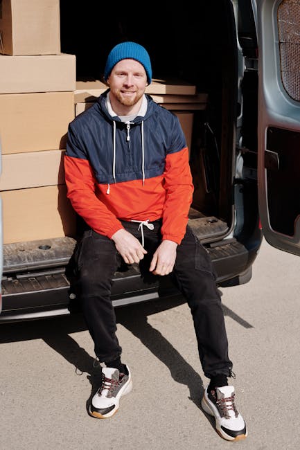 A man dressed in a blue beanie, navy and red windbreaker jacket, black pants, and white sneakers is sitting on the edge of an open van's loading area, surrounded by several large cardboard boxes. The boxes are stacked neatly on the vehicle's floor and behind him, indicating packing for a house relocation. The van, parked outdoors on a paved surface, has its rear doors wide open, revealing more boxes inside along with packing materials such as plastic wrap and fabric covers. The man is smiling, with one hand resting on his knee and the other on a box, suggesting he is taking a break during the furniture transport and packing process. The environment appears bright, likely daytime, with natural light illuminating the scene. This image captures a moment of loading and packing for a professional removals service, exemplifying careful handling of belongings during home relocation, as provided by Man and Van West Hampstead.