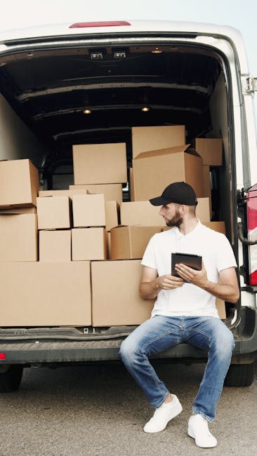 Two individuals, a man and a woman, are standing behind an open van, which is parked on a paved road in a suburban area under a partly cloudy sky. The van's large rear doors are fully open, revealing an interior filled with neatly stacked cardboard boxes of various sizes, some sealed with packing tape and others open, ready for loading or unloading. The man, dressed in a white t-shirt and jeans, is looking at his phone, while the woman, wearing a white cap, black sleeveless top, and denim skirt, is holding a clipboard or notes, possibly coordinating the move. The scene depicts a home relocation or packing process, with the boxes positioned in the van's cargo area, close to a pavement and grassy verge. Behind them, a street lamp and residential area are visible, indicating a typical neighbourhood setting. This image illustrates furniture transport and packing during a professional move facilitated by a house removals service such as Man and Van West Hampstead.