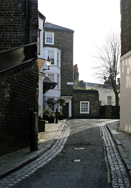 A narrow residential street during daytime in West Hampstead, featuring a cobblestone pathway bordered by a brick wall on the left and modern white and grey brick buildings with bay windows on the right. The street curves gently and is mostly empty, with a few leafless trees visible in the background. There are black bollards and lamp posts along the pavement, indicating a pedestrian-friendly area. This setting illustrates the typical environment where professional house removals, such as those offered by Man and Van West Hampstead, might take place, including residential streets suitable for home relocation or furniture transport during packing and moving processes.