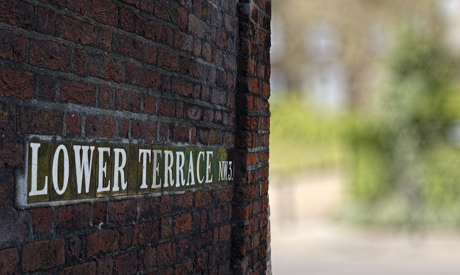 Close-up view of a brick exterior wall displaying a street sign that reads 'Lower Terrace NW3', with the sign mounted on dark red bricks and featuring white and green text. In the background, the image shows a blurred outdoor scene with trees and greenery, suggesting a residential area of West Hampstead. The lighting indicates daytime, with natural light illuminating the brick surface. This visual context relates to house removals and relocating services provided by Man and Van West Hampstead, highlighting the typical environment involved in home relocation and furniture transport tasks within the West End Lane area, supporting the theme of packing, loading, and moving processes.